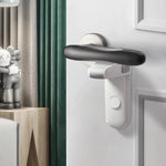 Ett vitt barnlås är monterat under ett svart dörrhandtag på en vit dörr. Låset är aktiverat och hindrar handtaget från att tryckas ned. I bakgrunden syns ett rum med gardin och en hylla med dekorationer.