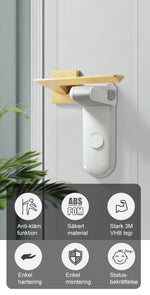Ett barnlås för dörrhandtag sitter monterat på en vit dörr med ett guldigt dörrhandtag. Låset är i vitt och grått och blockerar handtaget från att tryckas ner. Längst ned i bilden visas ikoner med text som beskriver funktioner: anti-klämfunktion, säkert ABS/POM-material, stark 3M VHB-tejp, enkel hantering, enkel montering och statusbekräftelse.