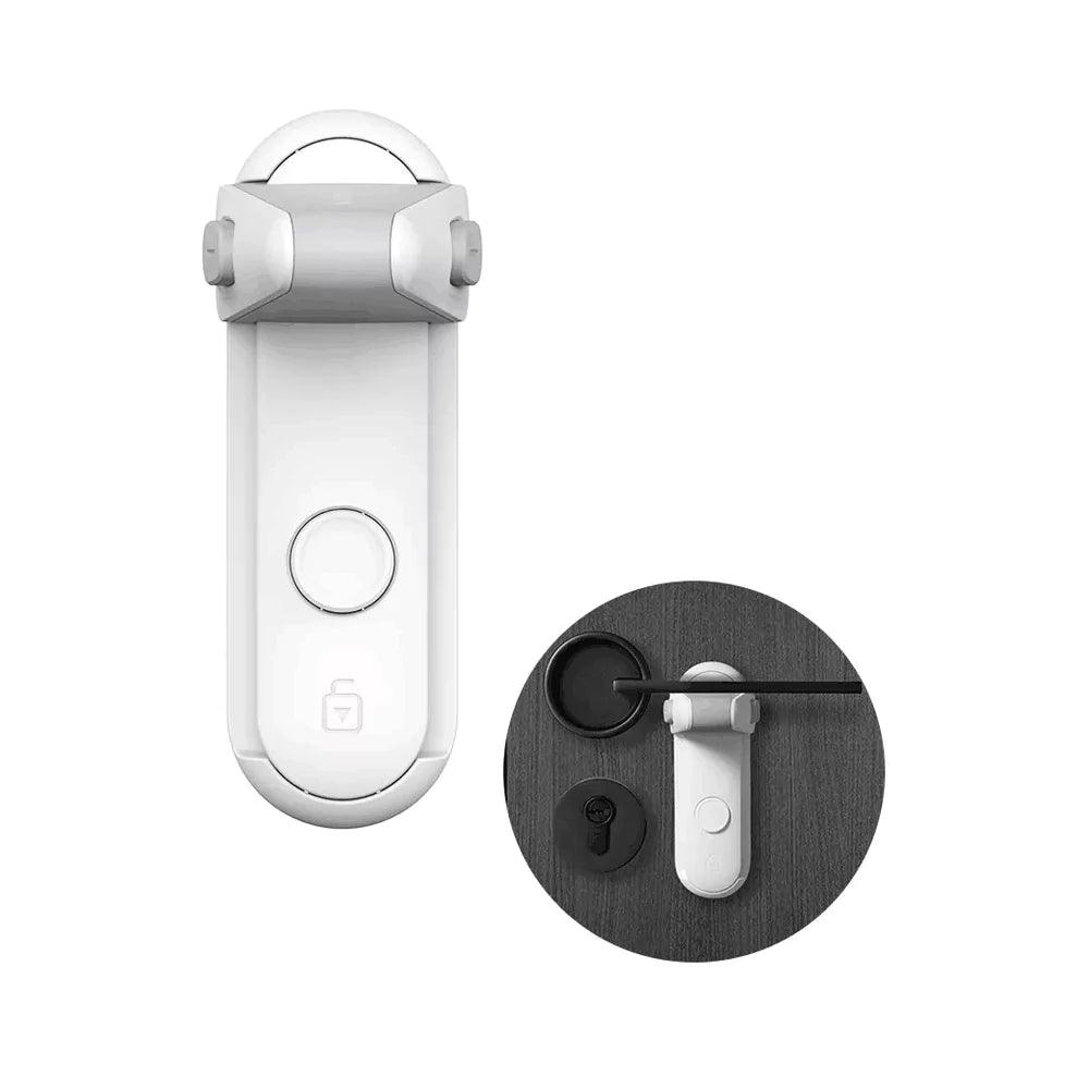 Vitt barnlås för dörrhandtag sedd framifrån, med ett rundat låsblockerande skydd upptill och en knapp på framsidan. I en mindre cirkel bredvid visas hur låset används på en mörk dörr där ett svart dörrhandtag hindras från att tryckas ned av låset.