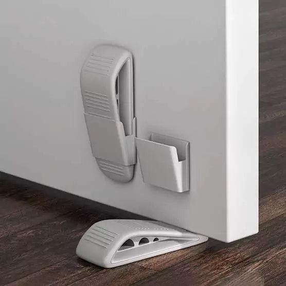 Grå dörrkil placerad under en vit dörr, med ett matchande väggfäste i plast monterat på dörrens insida för praktisk förvaring av kilen när den inte används.