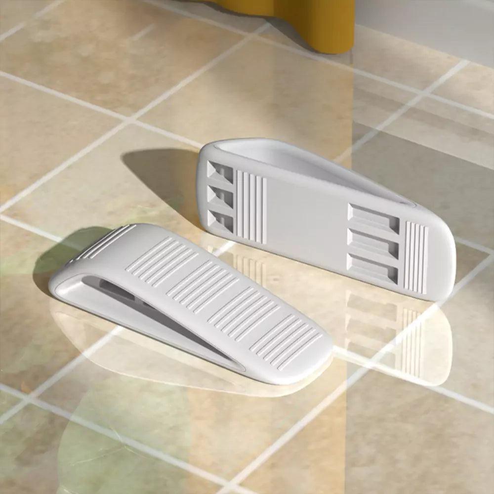 Två vita dörrkilar ligger på ett blankt, beige kakelgolv. Den ena dörrkilen ligger med ovansidan upp och visar ett räfflat halkskydd, medan den andra ligger på sidan och visar undersidans greppmönster.