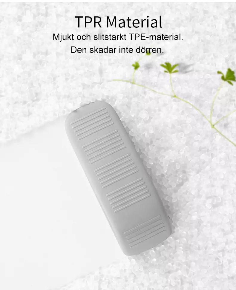 En grå dörrkil av TPE-material ligger på ett underlag av vita plastpellets, med några små gröna växtblad bredvid. Text ovanför beskriver att materialet är mjukt, slitstarkt och inte skadar dörren.