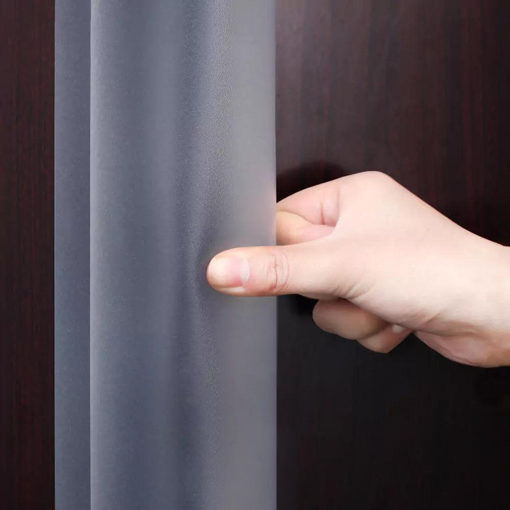 Hand som trycker på ett flexibelt, genomskinligt dörrklämskydd monterat på dörrens bakkant.