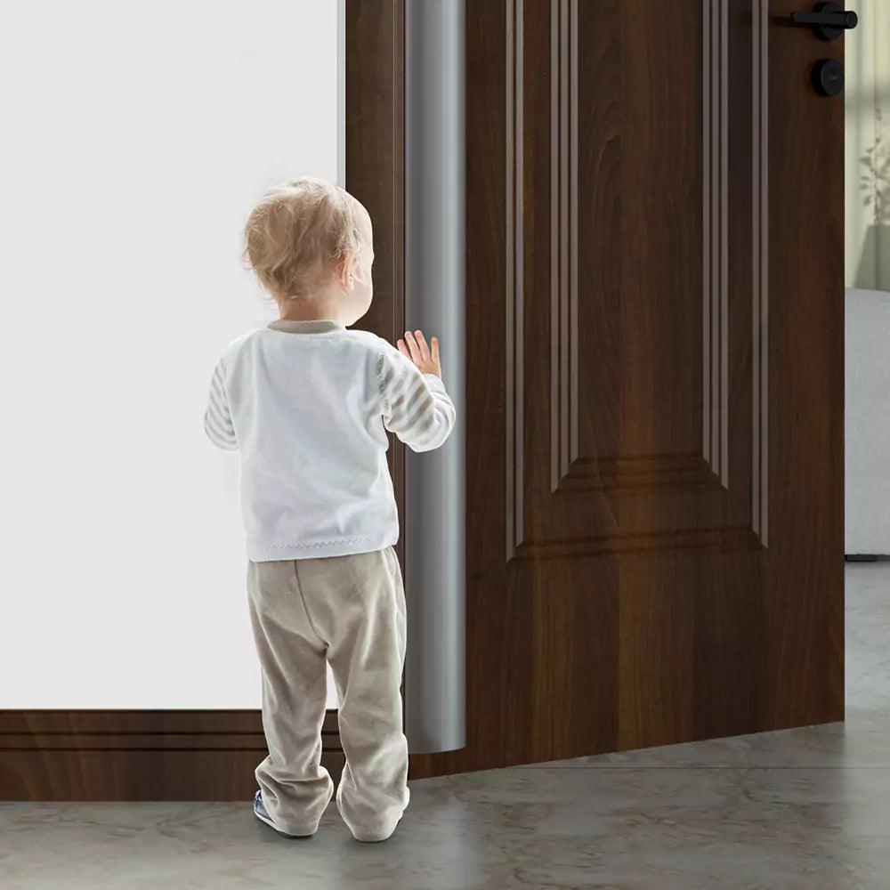 Litet barn som står vid en öppen dörr med ett monterat genomskinligt klämskydd på dörrens bakkant för att förhindra klämskador.