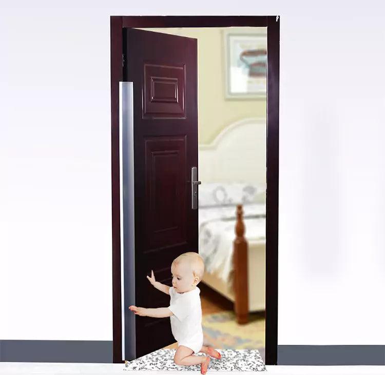 Litet barn som sitter på golvet vid en halvöppen dörr med ett monterat genomskinligt klämskydd längs dörrens bakkant för att förhindra klämskador.
