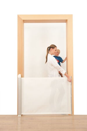 En vuxen person drar ut en vit, utdragbar säkerhetsgrind över en dörröppning. Bakom grinden står ett litet barn och tittar fram. Grinden är monterad mellan dörrkarmen och täcker den nedre delen av öppningen.