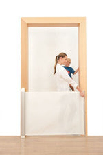 En vuxen person drar ut en vit, utdragbar säkerhetsgrind över en dörröppning. Bakom grinden står ett litet barn och tittar fram. Grinden är monterad mellan dörrkarmen och täcker den nedre delen av öppningen.