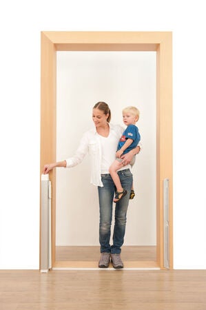 En vuxen person står i en dörröppning med ett litet barn på armen och räcker med den andra handen mot den infällda, hoprullade säkerhetsgrinden som sitter monterad på dörrkarmen.