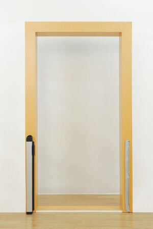 En tom dörröppning med en säkerhetsgrind monterad på vänster sida och en smal mottagare på höger sida. Själva grinden är infälld och inte utdragen, och ramen är i ljust trä mot en vit vägg.