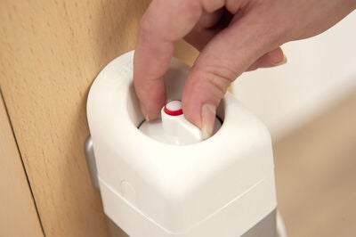 En hand trycker ned en vit knapp med en röd markering på ovansidan av en säkerhetsgrind monterad mot en ljus träyta.